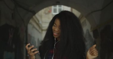 Gorgeous african girl with natural make-up and curly hair is listening to music in earphones and happily dancing in the street. Emotional portrait