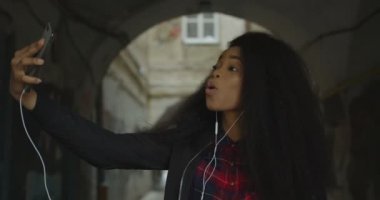 Side view of the attractive pretty african girl making funny faces while taking selfies via the mobile phone outdoor. She is making surprised face and showing tongue