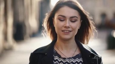 Front view of the young charming girl with natural make up is lovely smiling while walking along the sunny urban street and wind is moving her hair..