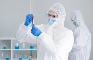 Virüs koruyucu giysiler içindeki epidemiyolojik araştırmacılar vida kapaklı test tüpündeki hastaların kan örneklerine bakıyorlar. Coronavirus hastalığı 2019 (COVID-19) bir laboratuvarda test süreci.