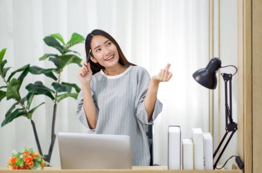 Genç Asyalı kadın kalemle şakağına dokunurken parmağını şıklatıyor. Üniversiteden bir ders kitabıyla evde çalışmanın tadını çıkar..