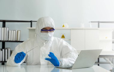 Virüse karşı koruyucu giysi giyen epidemiyolojik araştırmacılar dizüstü bilgisayar ekranına bakarken İğne ve Kimyasal Petri Çanağı 'nı tutuyorlar. Bir laboratuarda COVID-19 test süreci.