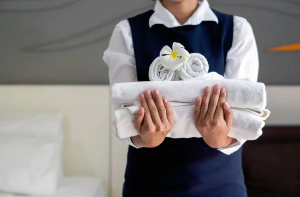 Genç otel hizmetçisinin elinde beyaz çiçekli yakın plan beyaz havlu. Mavi üniformalı personel otel misafirleri için oda hazırlıyor..