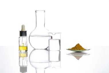 Beyaz laboratuvar masasında yakın çekim kimyasal malzeme. Damla şişesinde sarı kozmetik sıvısı düz dipte kristal berrak sıvı ve Kimyasal Saat Bardağında Organik Curcuma tozu.