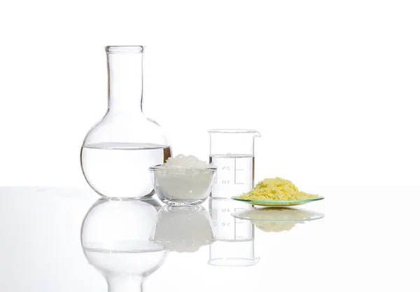Beyaz laboratuvar masasında yakın çekim kimyasal malzeme. Plastik taban şişesinde kristal berrak sıvı, beyaz pullu kimyasal ve kimyasal saat bardağının yanındaki Sodyum sülfür taneciği..