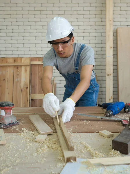 Toz geçirmez gözlüklü Asyalı marangoz bir tahta parçasına çizgi çekiyor ve bölümlere ayırmaya hazırlanıyor. Atölyede sabah çalışma ortamı.