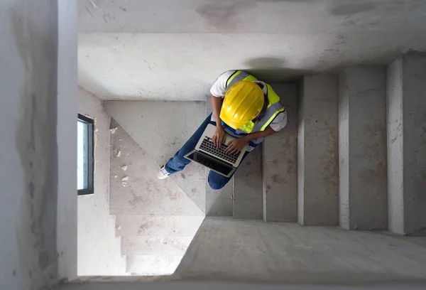 Engineer in hardhat and safety vest typing on laptop keyboard with blank screen while sit on a stairway. Work environment of engineers at the construction site of housing projects. Top View