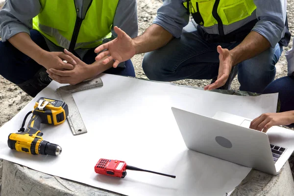 Real estate construction project meeting on a table made of concrete mixer bucket. Human hands, laptop computer, walkie-talkie, electric drill and engineer's square are on white paper.