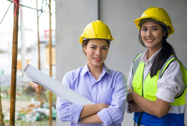 Young woman in construction helmet and floor plan stand with arms crossed at the construction site of housing projects. Asian engineer in a safety vest stand smiling beside her.