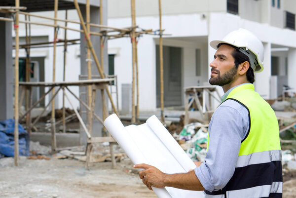 Young engineer in a construction helmet and safety vest holding a floor plan while looking at the progress of real estate projects. Day time work safety checks.