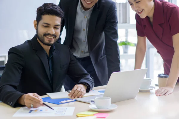 Young wavy hair, moustache and beard businessman in black suit meeting with colleague in modern office. Laptop computer, coffee,  document, graph, chart are on wooden table.