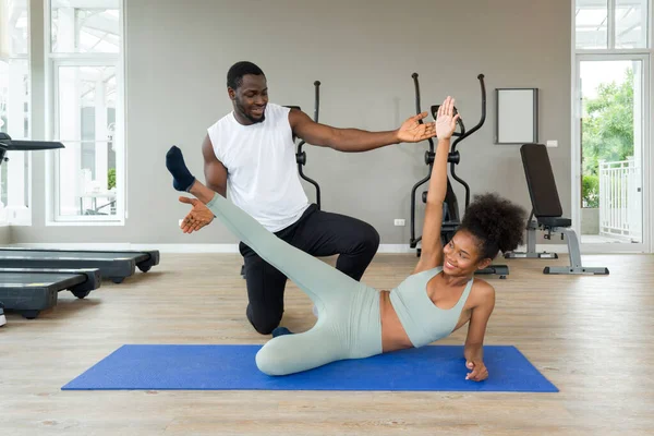 Short curly black hair coach with moustache and beard teach young woman in sportswear how to do side plank crunch. Cardio machines are on the background at the gym.