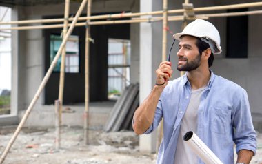Emlak projelerinin gidişatına bakarken elinde Walkie-Talkie tutan inşaat kasklı genç bir mühendis. İnşaat alanında ahşaptan yapılmış iskeleler var..
