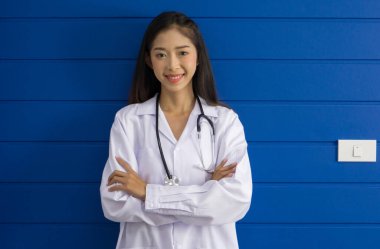 Young asian doctor in white gown and stethoscope stand with arm cross in front of blue wall.