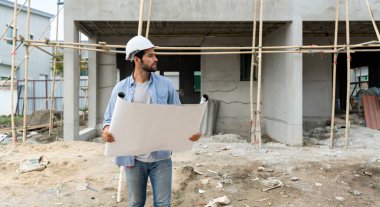 Emlak projelerinin gidişatına bakarken elinde kat planı tutan miğferli genç bir mühendis. İnşaat alanında ahşap ve fiber beton çatı kiremitlerinden yapılmış iskeleler var..