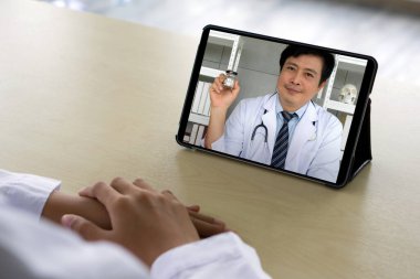 Beyaz önlüklü eczacının arka planda oturduğu masada Asyalı doktorla tıbbi kapsül hakkında tablet bilgisayarda webcam konferansı var. Sağlık ve Tıp Konsepti.