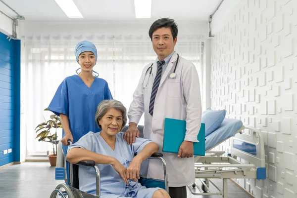 Senior female patient and doctor. Young nurse push a wheelchair of the elderly patient while asian doctor with stethoscope and gown stood beside.