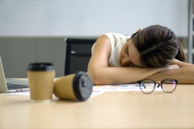 Young asian employee take a nap in the office on a table with coffee cup, document, eyeglass and laptop computer.