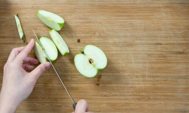 Closeup hand holding knife cutting green apple on a wooden chop board. Top View.
