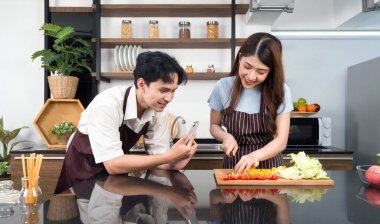 Asian couple spend time together in the kitchen. Young woman in apron cooking salad dish while his boyfriend recording vlog video for social blogger. Modern lifestyle  people relationship and activity