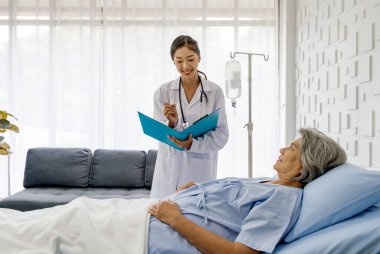 Young asian doctor in white gown with stethoscope and document folder, examine the symptom of the elderly patient in the recovery room. Healthcare and medicine concept