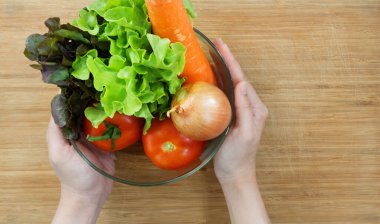 Closeup hand holding a glass bowl with a variety of vegetables on wooden table. Top View. Onion, carrot, tomato, Red and Green Oak Lettuce