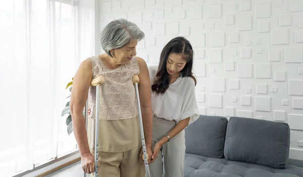 Young asian woman help her grandma walk by using axilla crutches. Senior gray hair woman exercise and practice walking at home.