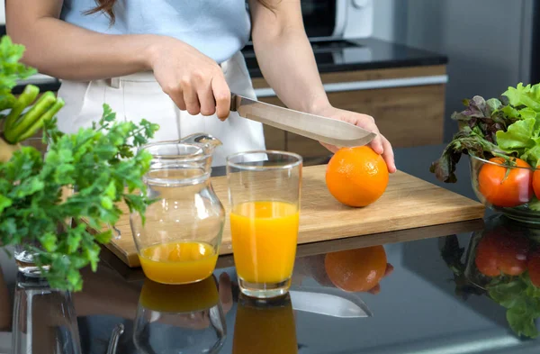 Closeup hand holding knife cutting orange fruit on a wooden chop board. Jar with mixed fruit juice  and glass bowl with a variety of fruits and vegetables is placed on the kitchen counter.