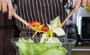 Ev hanımı önlük giymiş, sebzeleri ahşap bir kepçeyle salata kasesine karıştırmış. Domates, dolmalık biber ve marul. Kapat.