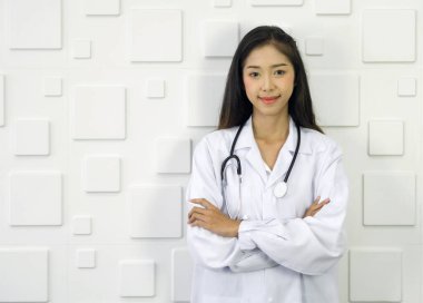 Beyaz önlüklü ve steteskoplu genç Asyalı doktor beyaz dikdörtgen desenli seramik duvarın önünde kolları çapraz duruyor..