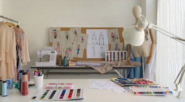 Moda tasarımcısı odası. Dikiş makinesi ve masadaki çeşitli dikiş malzemeleri. Ayakta duran mankenler, renkli kumaşlar ve masanın yanında çeşitli elbise rafları..