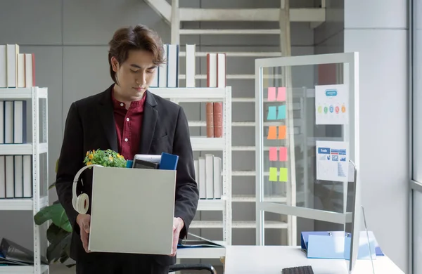 Young asian employee in black suit feel sad, stressed and unhappy face holding paper box of belongings collected from office in the get fired day. Idea for unemployed person, work crisis.