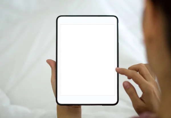 Back view of woman typing on blank screen tablet computer while sit relax on bed with white duvet. Close up.