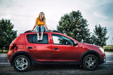 Bir kadın arabanın çatısında oturur ve yola bakar..