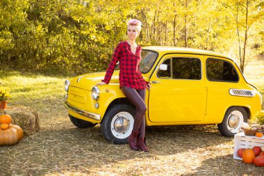 Sarı retro arabanın yanında kısa saçlı bir kadın sonbahar parkında altın ağaçlarla arka planda.