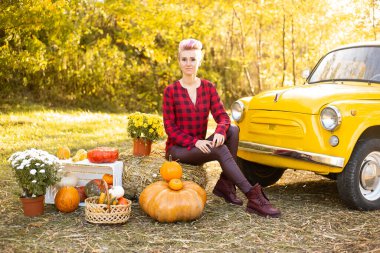 Sarı arabanın yanında gülümseyen bir kadın çiçekler ve balkabaklarıyla sonbahar parkında altın ağaçlarla birlikte.