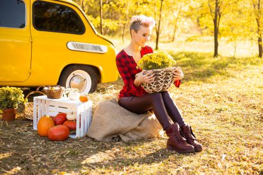 Sonbahar parkında sarı arabanın yanında çiçekli ve balkabaklı gülümseyen güzel bir kadın.