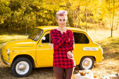 Sonbahar parkında arka planda altın ağaçlarla sarı retro arabanın yanında kısa saçlı şık hippi bir kadın.