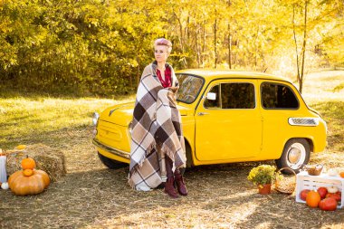 Sarı retro arabanın yanındaki battaniyeye sarılmış bir kadın. Sonbahar çiçeklerinin arka planında altın ağaçlarla birlikte.