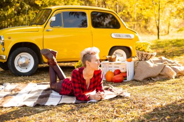 Sonbahar parkında rahatlama, güz parkında battaniyeye uzanmış sıradan giyinmiş mutlu hippi kadın sarı arabanın, çiçeklerin ve balkabaklarının yanında.