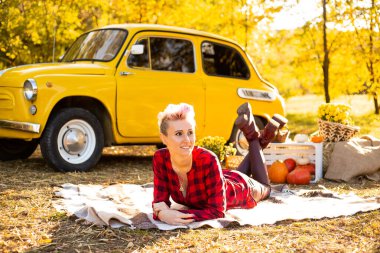 Sonbahar parkında, arka planda, sarı arabanın, çiçeklerin ve balkabaklarının yanında, kısa saçlı, güler yüzlü hippi kadın.