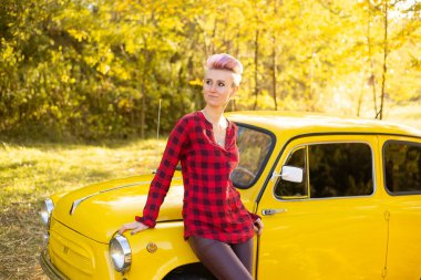 Sonbahar parkında sarı arabanın yanında kısa saçlı, altın ağaçlı bir hippi kadın.
