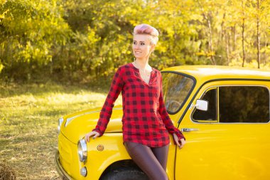 Sonbahar parkında sarı arabanın yanında kısa saçlı, altın ağaçlı bir kadın.