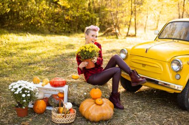 Sonbahar parkında sarı arabanın yanında çiçekli ve balkabaklı mutlu bir kadın.