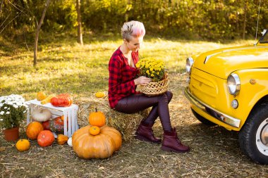 Sonbahar parkında çiçekli ve balkabaklı bir kadın sarı arabanın yanında.