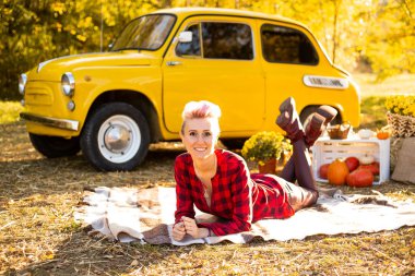 Sonbahar parkında, arka planda, sarı arabanın, çiçeklerin ve balkabaklarının yanında battaniyeye uzanmış bir kadın.