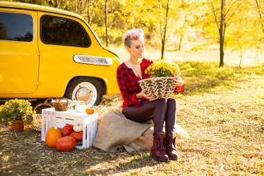 Sonbahar parkında sarı arabanın yanında çiçekli ve balkabaklı mutlu bir kadın.