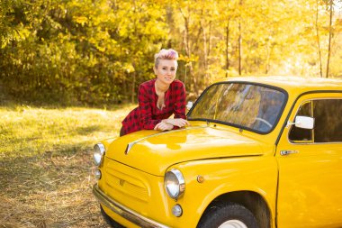 Sonbahar Park arka planında, sarı retro arabanın yanında kısa saçlı, altın ağaçlı hippi bir kadın.
