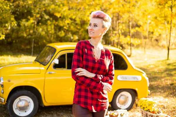 Sonbahar parkında arka planda altın ağaçlarla sarı retro arabanın yanında kısa saçlı mutlu şık bir kadın.
