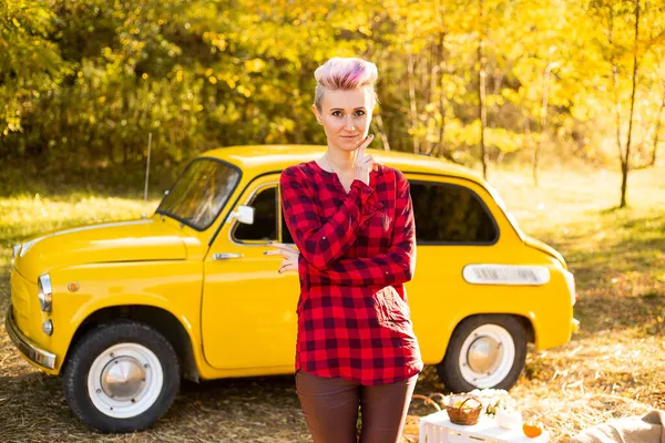Sonbahar parkında arka planda altın ağaçlarla sarı retro arabanın yanında kısa saçlı şık hippi bir kadın.